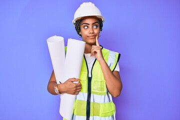 Young african american woman with braids wearing safety helmet holding blueprints serious face thinking about question with hand on chin, thoughtful about confusing idea