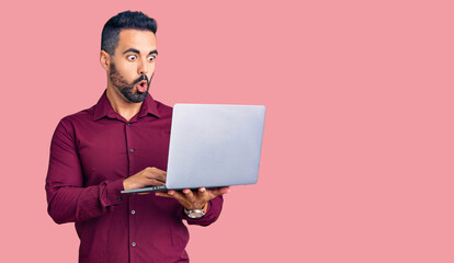 Young hispanic man holding laptop scared and amazed with open mouth for surprise, disbelief face