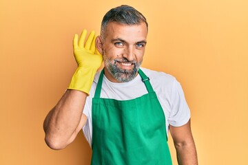 Middle age handsome man wearing cleaner apron and gloves smiling with hand over ear listening and hearing to rumor or gossip. deafness concept.