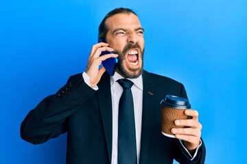 Attractive business man with long hair and beard using smartphone and drinking a cup of coffee angry and mad screaming frustrated and furious, shouting with anger. rage and aggressive concept.