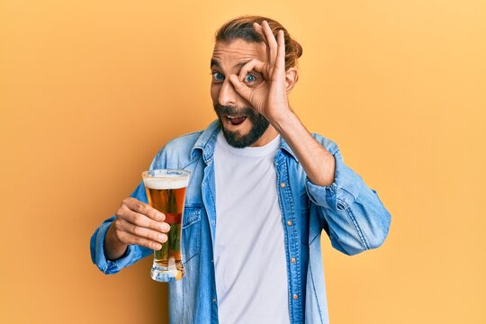 Attractive man with long hair and beard drinking a pint of beer smiling happy doing ok sign with hand on eye looking through fingers