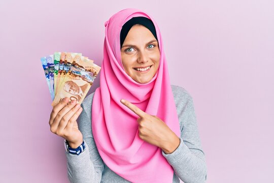 Young caucasian woman wearing islamic hijab holding canadian dollars banknotes smiling happy pointing with hand and finger