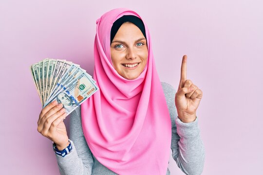Young caucasian woman wearing islamic hijab holding dollars banknotes smiling with an idea or question pointing finger with happy face, number one