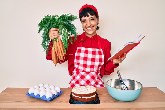 Beautiful brunettte woman cooking carrot cake reading recipes book smiling with a happy and cool smile on face. showing teeth.