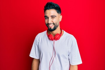 Young man with beard listening to music using headphones winking looking at the camera with sexy expression, cheerful and happy face.