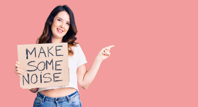 Beautiful young brunette woman holding make some noise banner smiling happy pointing with hand and finger to the side