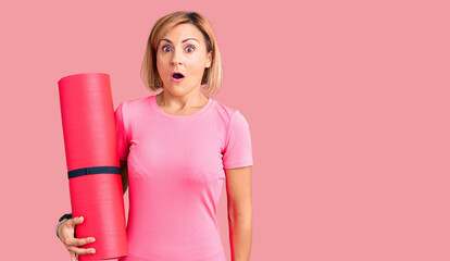 Young blonde woman holding yoga mat scared and amazed with open mouth for surprise, disbelief face