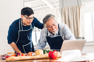 Portrait of happy love asian family senior mature father and young adult son having fun cooking together and looking for recipe on Internet with laptop computer to prepare the yummy eating lunch 