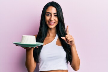 Beautiful hispanic woman eating fresh white cheese smiling with an idea or question pointing finger with happy face, number one