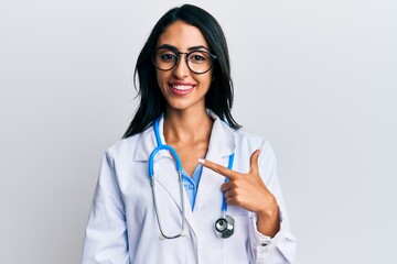 Beautiful hispanic woman wearing doctor uniform and stethoscope smiling cheerful pointing with hand...