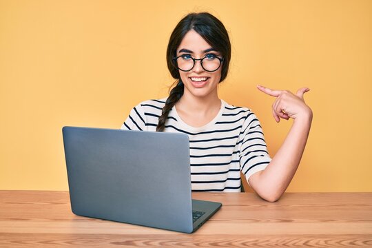 Brunette teenager girl working using computer laptop pointing finger to one self smiling happy and proud