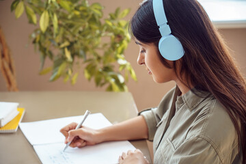 Indian woman student wearing headphones writing homework in notebook studying at home listening...