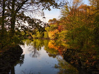 Looking Down River on Brisk October Day with Beautifully Colored Trees with Leaves in Orange, Yellow, Red and Green Reflected in Water with Bright Blue Sky in Background