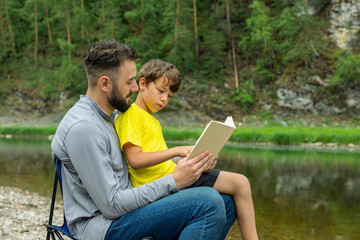 Fototapeta premium smiling man and young boy reading book on nature. Family holiday and togetherness.