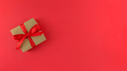 Gift box wrapped in a craft paper with red ribbon and bow on red background. Monochrome festive flat lay with copy space.