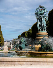 A statue of people holding a globe and horses splashing in the water of the fountain greets visitors to Luxembourg Gardens in Paris, France, with the palace in the distance on a sunny summer day.