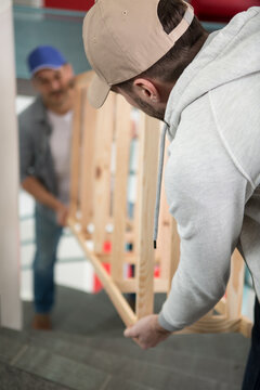 Two Male Contractors Carrying Wooden Frame Up Some Stairs