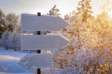 Empty white track pointers, guidepost in sunlight against winter nature background. Directional arrow signs on wooden pole in snowy forest.