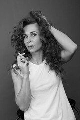Obraz premium A beautiful brunette girl with long curly hair in a white T-shirt smokes in the studio on a gray background, bw