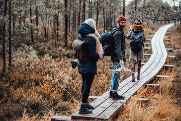 Group of three friends guy and two girls going through wooden path following each other in autumn lovely forest.
