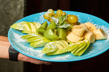 A man holds a blue plate with sliced apples, bananas, kiwi and grapes