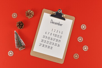 Christmas flat lay with clipboard with calendar sheet for month December surrounded by small Christmas trees, pine cones, snowballs and snowflake ornaments on red background
