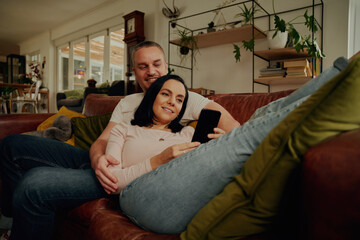 Obraz premium Smiling young woman using phone while relaxing with husband on couch at home