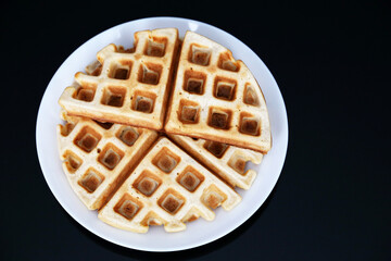 Belgian waffles in white plate on black glass table, top view. Healthy breakfast, pieces of freshly baked classic wafer