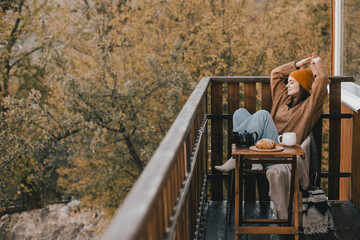 Young woman in knitted sweater and hat drinking tea and eating fresh croissants on cozy balcony of...