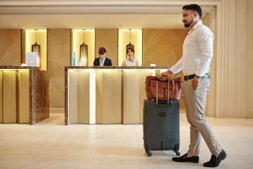 Handsome elegant man with briefcase and suitcase entering lobby of expensive hotel
