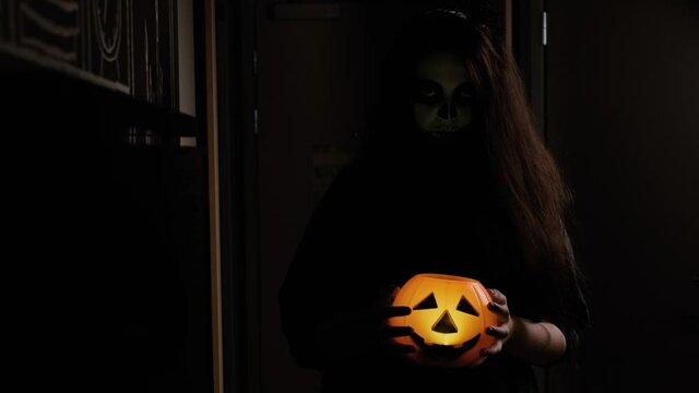 A girl with long hair, black clothes and zombie make-up walks down a dark corridor, holding a glowing orange pumpkin head in her hands. Holiday attribute and symbol. Halloween Eve. October holiday.