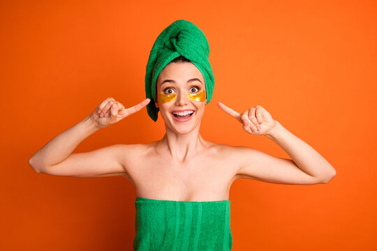 Photo portrait of woman pointing fingers at golden eye patches wearing green bath towel isolated on vivid orange colored background