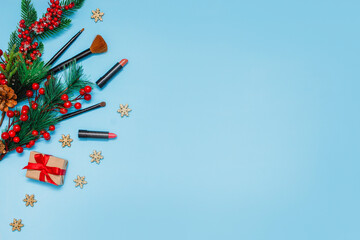 red lipsticks, makeup brushes and a gift lie on a blue background next to spruce branches with red berries. cosmetics for festive makeup for Christmas. Winter sales of cosmetics. Copy space, top view