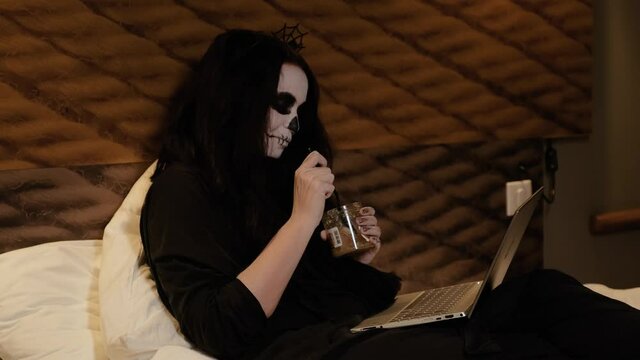 A girl with long hair in the form of a zombie with appropriate makeup, reclining on the bed, eating brain jam from a jar with a spoon, looking at a laptop and laughing. Halloween Eve. Festive humor.