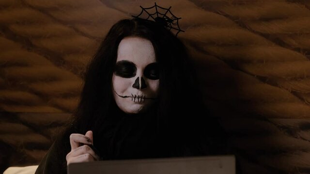 Close-up of the face of a girl with long hair in the image of a zombie with appropriate makeup, who sits on the bed, eats brain jam from a jar with a spoon, looks at a laptop and laughs. Halloween Eve
