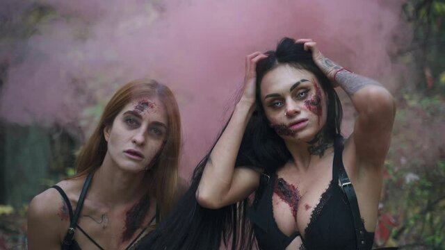 Two scary zombie women with wounds on their body and face stand against the background of pink smoke