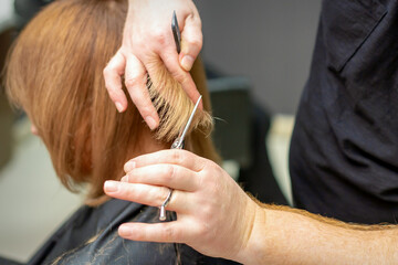 Back view of hairdresser cuts red or brown hair to young woman in beauty salon. Haircut in hair salon