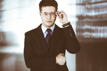 Young businessman wearing suit, with glasses, while standing in the office. Business success concept nowadays