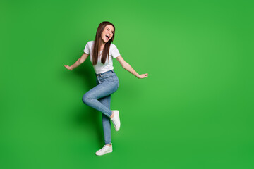 Full length photo of adorable dreamy young lady dancing wear jeans t-shirt footwear isolated on green background