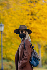 Style blonde in face mask and coat on autumn park with maples on background