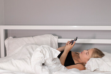 attractive blonde under the covers with white sheets surfing the internet on the phone. pretty people on isolation.