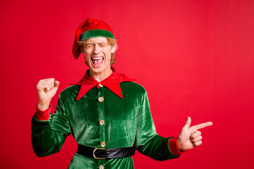 Photo of delighted elf guy point finger copyspace x-mas christmas ads raise fists isolated on red...
