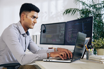 Pensive young Indian programmer sitting at office desk with two monitors and typing on laptop