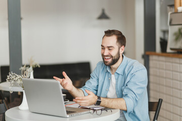 Smiling funny young man sitting alone at table in coffee shop cafe working or studying on laptop pc computer relaxing in restaurant during free time indoors. Freelance, mobile office business concept.