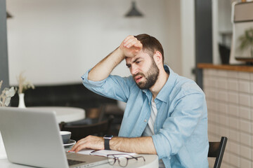Naklejka premium Sick tired exhausted young man sitting alone at table in coffee shop cafe restaurant indoor working or studying on laptop pc computer put hand on head. Freelance mobile office business concept.