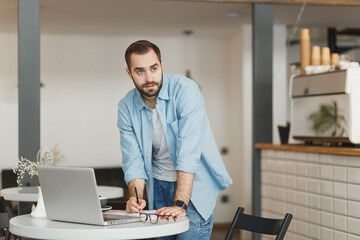 Fototapeta premium Confident handsome young man standing alone near table in coffee shop cafe restaurant indoors working or studying on laptop pc computer writing in notebook. Freelance mobile office business concept.