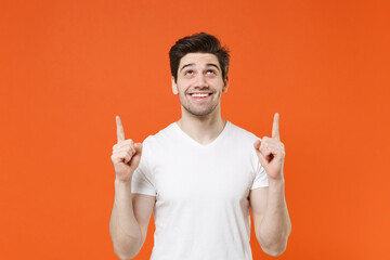Smiling attractive young man 20s wearing basic casual empty blank white t-shirt standing pointing index fingers up on mock up copy space isolated on bright orange colour background, studio portrait.