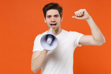 Dissatisfied young man 20s wearing basic casual empty blank white t-shirt standing screaming in megaphone clenching fist looking camera isolated on bright orange colour background studio portrait.