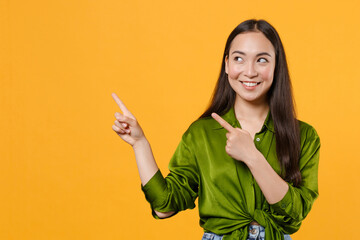 Smiling beautiful young brunette asian woman wearing basic green shirt standing pointing index fingers aside up on mock up copy space isolated on bright yellow colour background, studio portrait.