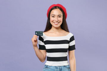 Beautiful attractive smiling young brunette asian woman 20s wearing striped t-shirt red beret standing hold credit bank card looking camera isolated on pastel violet colour background studio portrait.